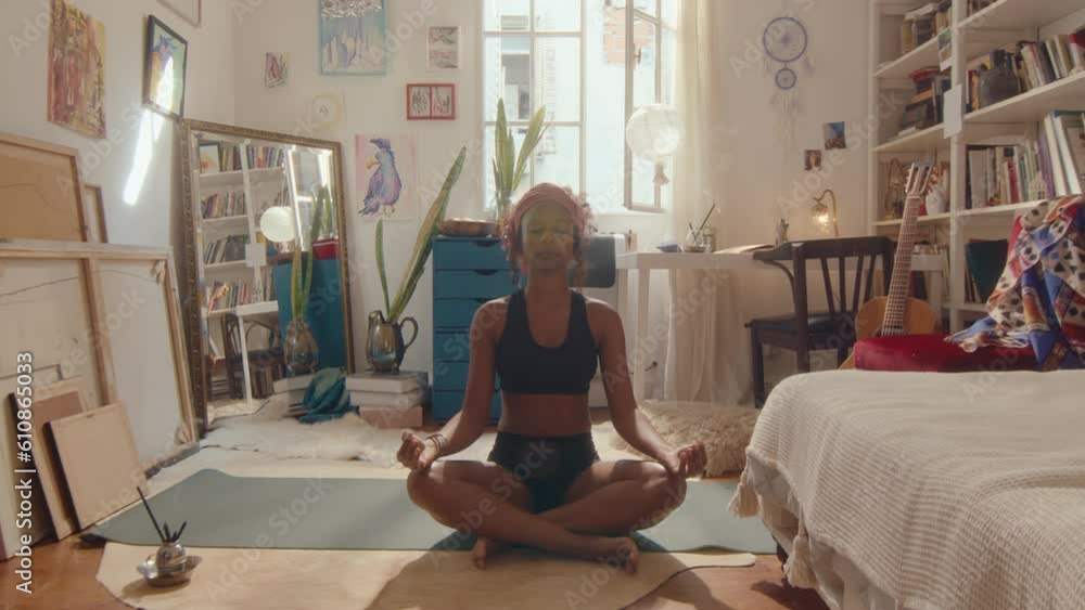 Young black girl sitting with eyes closed in lotus pose on mat, holding ...