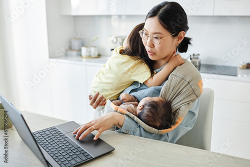 Asian young mother with two kids working online on laptop while sitting in the kitchen photo