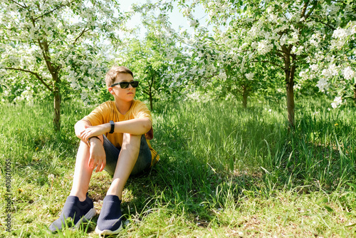 Boy in sunglasses sit in park under bloosom apple tree. Teen has rest after riding scooter on hot summer day