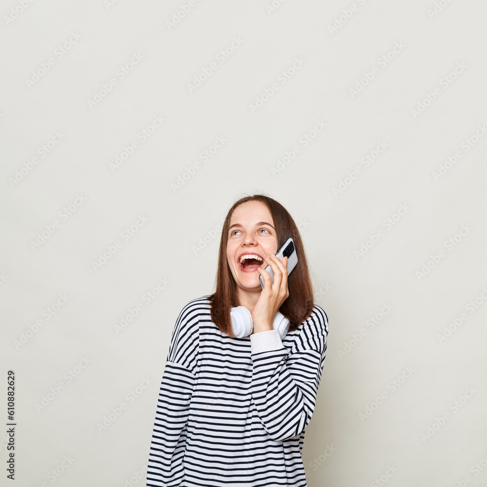 Laughing brown haired woman wearing striped shirt posing isolated over gray background talking on mobile phone copy space fore advertisement, mockup. Laughing brown haired woman wearing striped shirt posing isolated over gray background talking on mobile phone copy space fore advertisement, mockup.