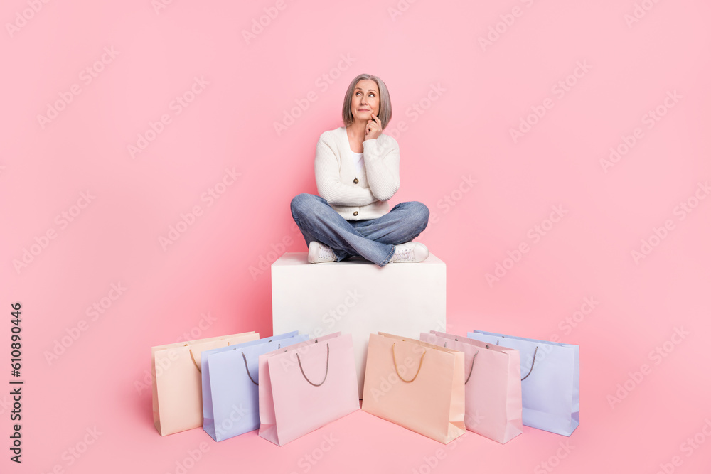 Full size photo of attractive senior woman sit cube thoughtful shopping bags wear trendy white clothes isolated on pink color background Full size photo of attractive senior woman sit cube thoughtful shopping bags wear trendy white clothes isolated on pink color background