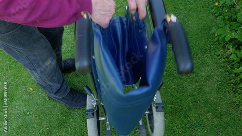 Closeup of a man’s hands grabbing two handles on the fabric seat of an empty, portable wheelchair, outside on grass, causing it to fold into a much narrower size, ready to be moved. photo