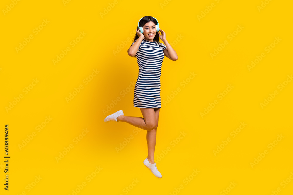 Full body length photo of korean beautiful girl new sony headphones quality music jumping wear striped dress isolated on yellow background Full body length photo of korean beautiful girl new sony headphones quality music jumping wear striped dress isolated on yellow background