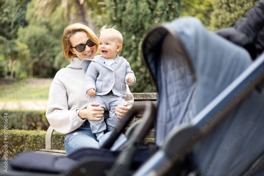 Mother sitting on bench in urban park, laughing cheerfully, holding her smiling infant baby boy child in her lap having baby stroller parked by their site.