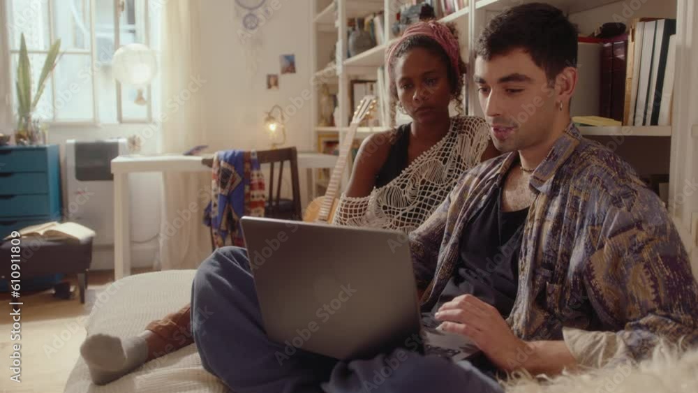 Young gen Z man using laptop and talking with beautiful black girlfriend, both sitting on bed in cozy sunlit apartment during the day Young gen Z man using laptop and talking with beautiful black girlfriend, both sitting on bed in cozy sunlit apartment during the day