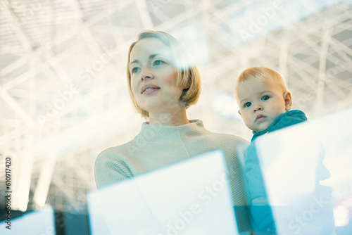 Thoughtful young mother looking trough window holding his infant baby boy child while waiting to board an airplane at airport terminal departure gates. Travel with baby concept photo