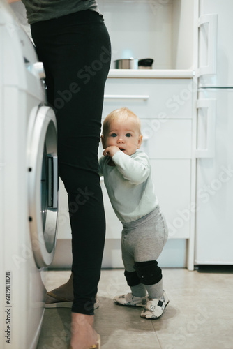Little infant baby boy child hiding between mothers legs demanding her attention while she is multitasking, trying to do some household chores in kitchen at home. Mother on maternity leave