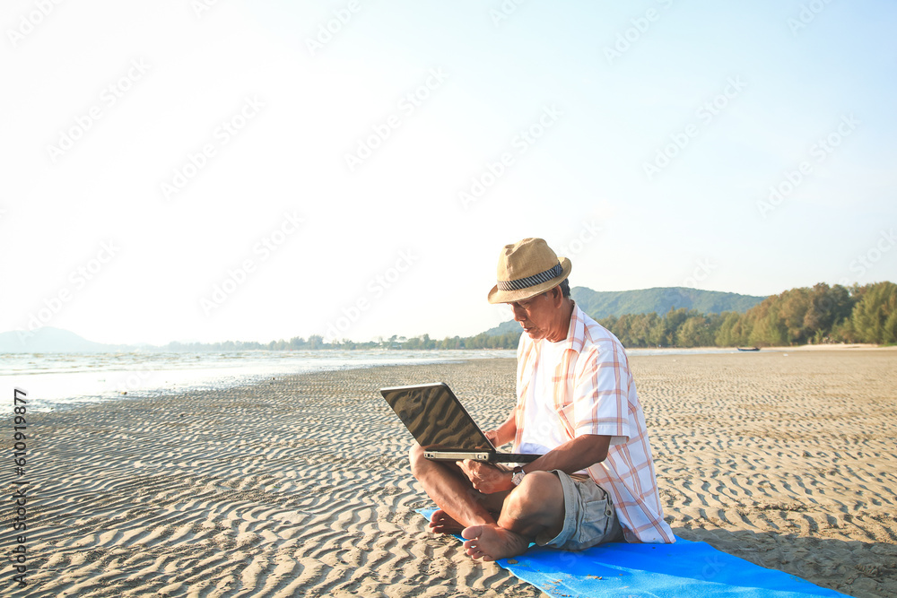 Asian elderly man sitting on the beach using laptop to work online. Living in retirement to be happy. aging society. copy space