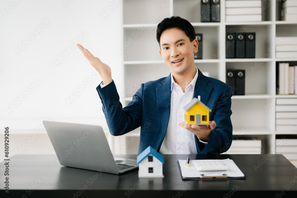 Young real estate agent worker working with laptop and tablet at table in modern office and small house beside it.. Young real estate agent worker working with laptop and tablet at table in modern office and small house beside it..