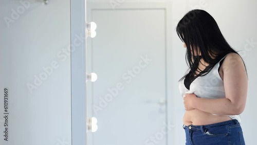 Chubby woman in t-shirt feeling insecure in own body touches fat folds on belly. Black-haired overweight lady looks at reflection on mirror at home