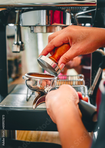 Hand of a barista holding a portafilter and a coffee tamper making an espresso coffee. Barista presses ground coffee using a tamper in a coffee shop