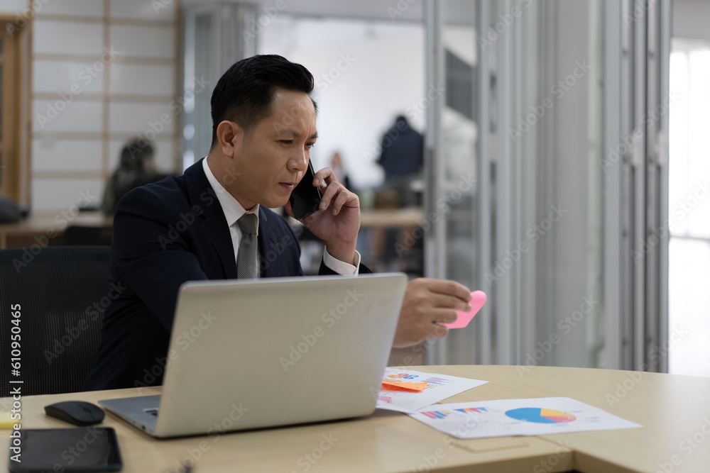 Businessman in suit talking on mobile phone in office. Portrait of professional Asian business people sitting at table with laptop computer using cellphone. Wireless smartphone is modern communication Businessman in suit talking on mobile phone in office. Portrait of professional Asian business people sitting at table with laptop computer using cellphone. Wireless smartphone is modern communication