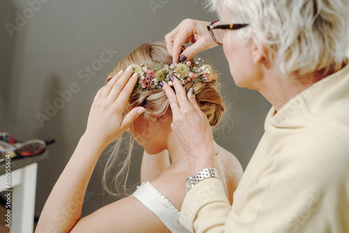 bride get ready with hair done from hairdresser with flowers and bouquet in updo hairstyle while hairstylist pin flower to head of woman
