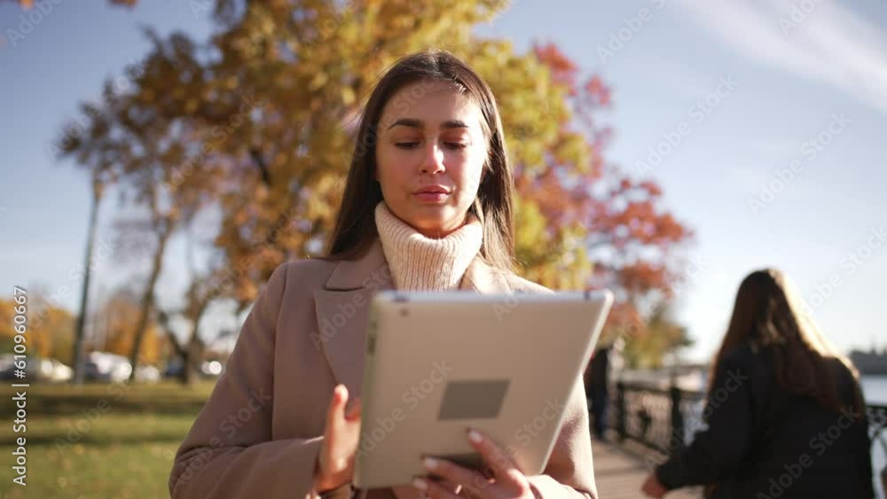 Portrait cute girl who walks on park in city center near embankment with water and uses digital tablet. Young woman looks at screen and typing. Online freelance work or text messages in communicate.