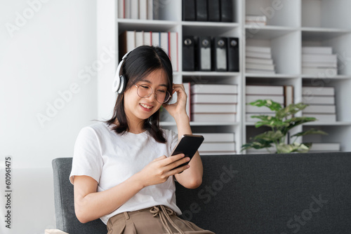 Young Asians woman listening to music on couch in living room at home. Happy Asia female using mobile smartphone, wearing headset and sitting on sofa