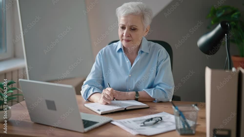 Woman sits office at table at workplace, writes on paper, looks at laptop screen talks on video call. Mature older gray-haired woman, professional accountant. Office worker. Positive businesswoman. Woman sits office at table at workplace, writes on paper, looks at laptop screen talks on video call. Mature older gray-haired woman, professional accountant. Office worker. Positive businesswoman.