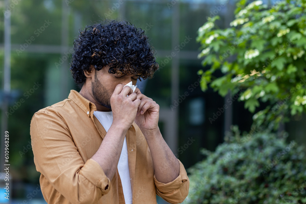 A young hispanic man stands on the street and wipes his nose with a tissue. He has a runny nose, flu, suffers from seasonal allergies A young hispanic man stands on the street and wipes his nose with a tissue. He has a runny nose, flu, suffers from seasonal allergies