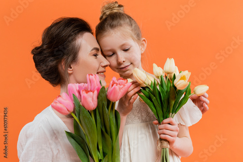 happy mother and daughter with tulips, young woman and girl holding flowers and posing on orange background, summer fashion, sun dresses, female bonding, family love