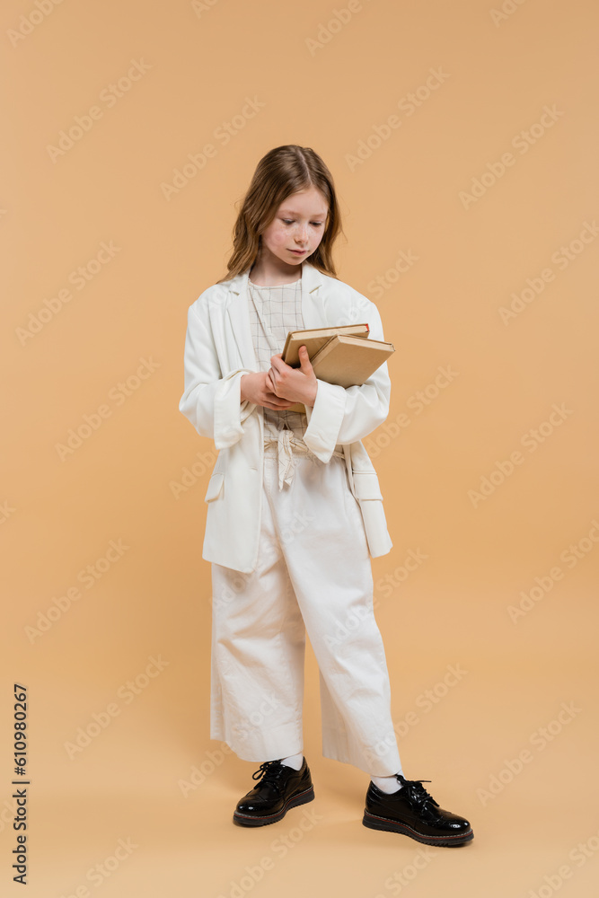 Fototapeta premium education concept, preteen girl in white suit holding books and standing on beige background, fashionable outfit, formal attire, back to school, preparing for school new year
