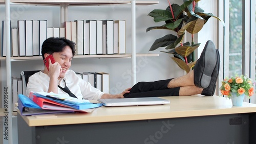 Young asian man putting feet on table in office while enjoy talking by smartphone take break from work. Happy businessman cellphone