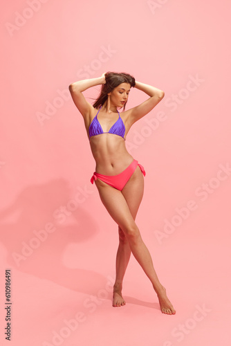 Full-length portrait of young, beautiful, brunette girl with slim, perfect body posing in swimsuit against pink studio background