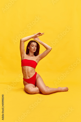 Attractive, brunette young girl with relief, fit, perfect body, in red swimsuit posing against yellow studio background © Lustre