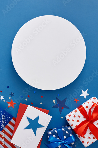 Celebration concept for America's Fourth of July holiday. Top view flat lay of gift boxes, postcard, american flag, event confetti on blue background with blank circle for text or greeting