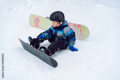 little boy sitting on snow putting his feet in snowboard bindings adjusting straps.