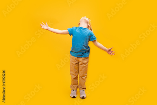 Little child standing over yellow background wearing in blue t-shirt and raised her hands up. Girl looks up