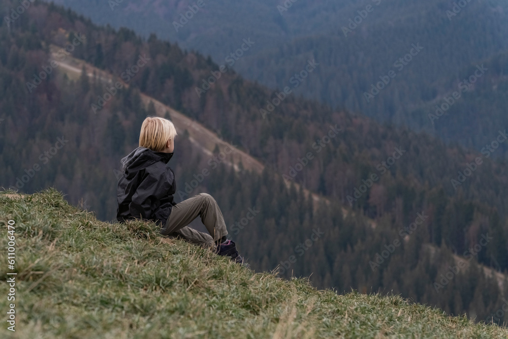 Blond boy sits at hillside and looks at the distance. Portrait of child in mountain in autumn.