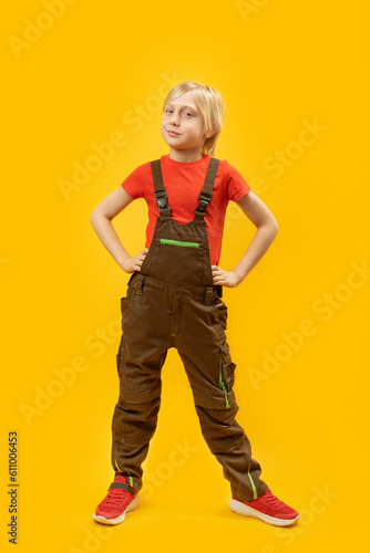 Vertical frame studio full-length portrait of teenage boy in overalls and red T-shirt on yellow background. Blond schoolboy 9-10 years old.
