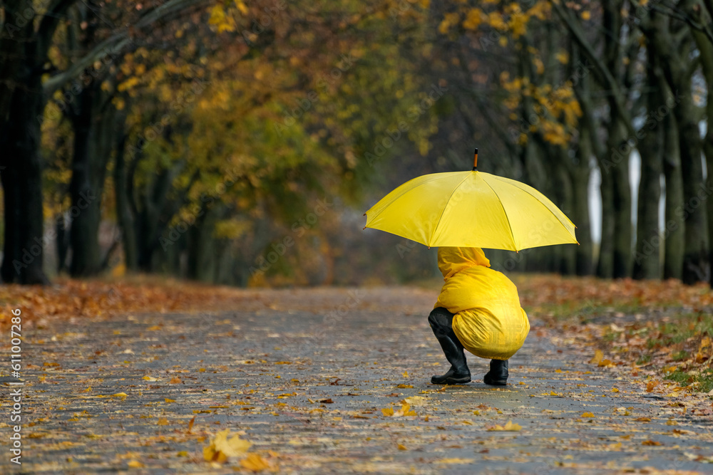 Child in yellow raincoat sits under yellow umbrella in an autumn park. Rainy fall day