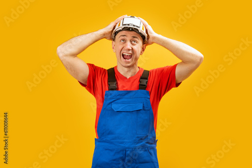 Young architect man with helmet with shocked facial expression, isolated yellow background . Worker or builder holding his head and screaming
