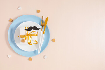 Father's Day concept. Flat lay top view of plate cutlery knife fork gift box, black mustache and hearts on pastel beige background with space for text or promotion and greeting message, banner