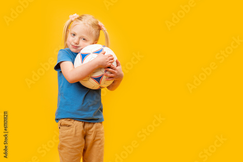 Little blonde girl holding soccer ball on yellow studio background. Copy space, mockup. Football for kids concept.