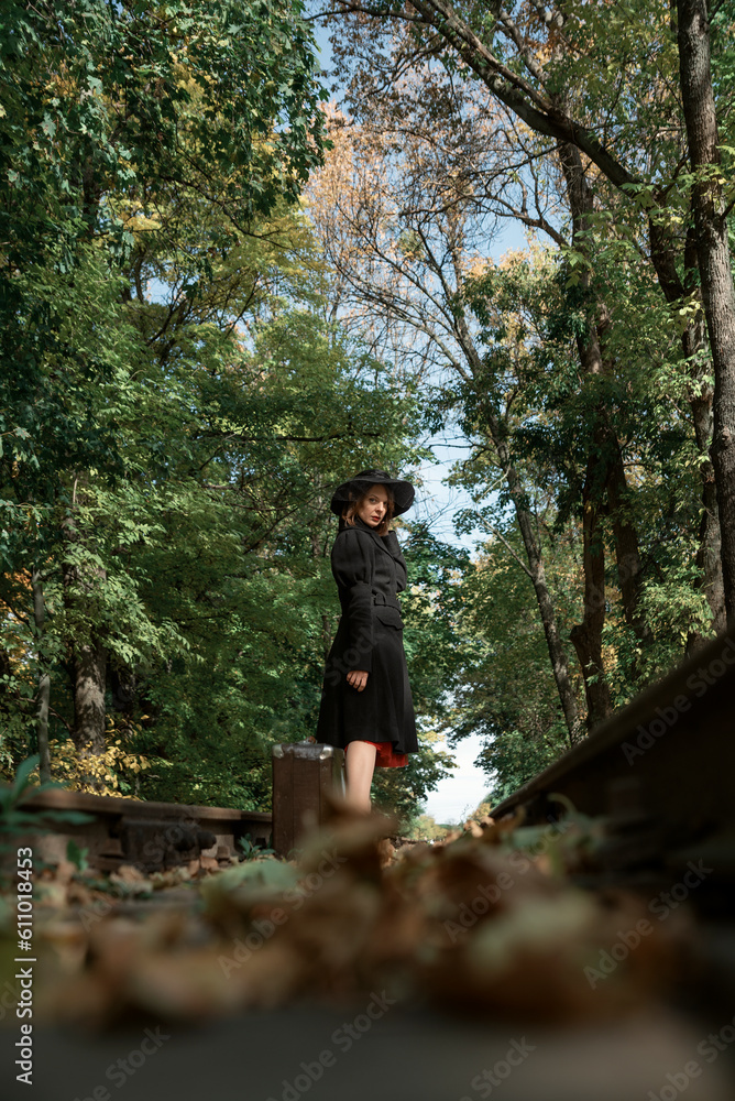 Beautiful woman in black coat, hat and with retro suitcase on rails on forest background. Vintage clothing. Bottom view Beautiful woman in black coat, hat and with retro suitcase on rails on forest background. Vintage clothing. Bottom view