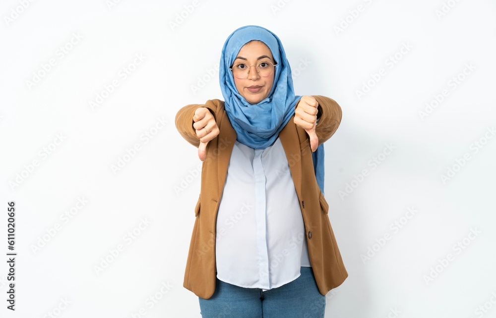 Young beautiful pregnant muslim woman wearing hijab in white studio being upset showing thumb down with two hands. Dislike concept.