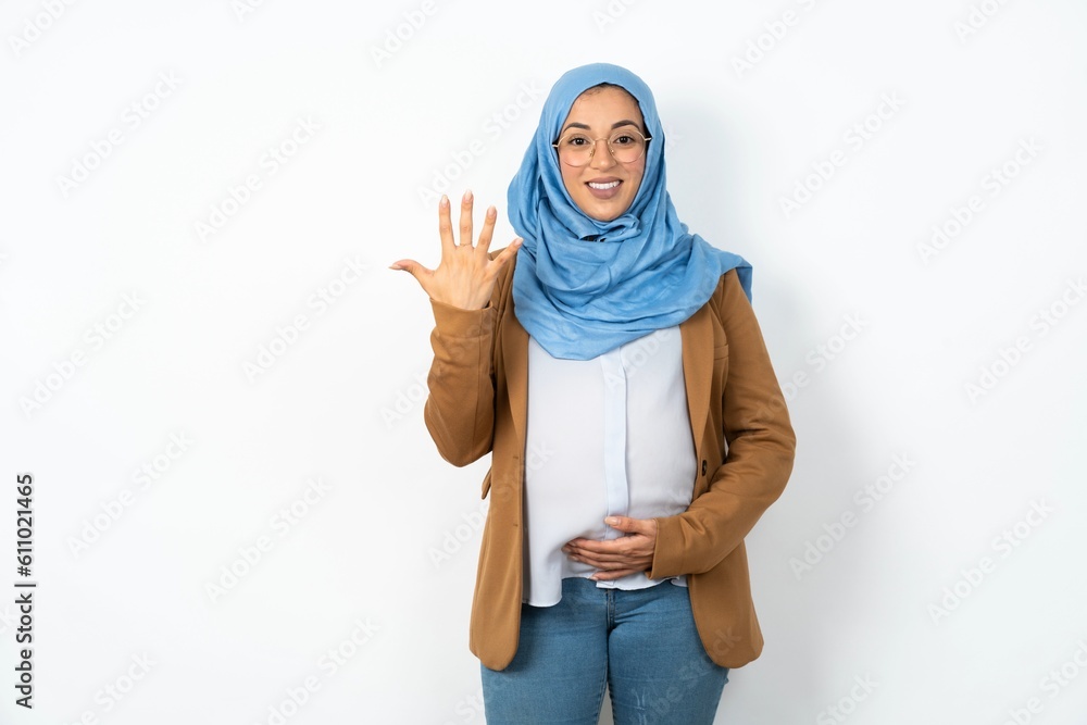 Young beautiful pregnant muslim woman wearing hijab over white background smiling and looking friendly, showing number five or fifth with hand forward, counting down Young beautiful pregnant muslim woman wearing hijab over white background smiling and looking friendly, showing number five or fifth with hand forward, counting down