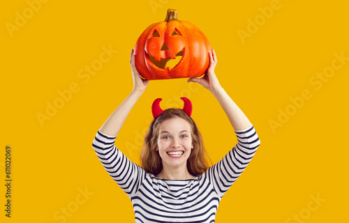 Happy woman holding up Halloween pumpkin. Cheerful beautiful young girl with devil horns standing isolated on yellow background  holding up carved pumpkin  looking at camera and smiling
