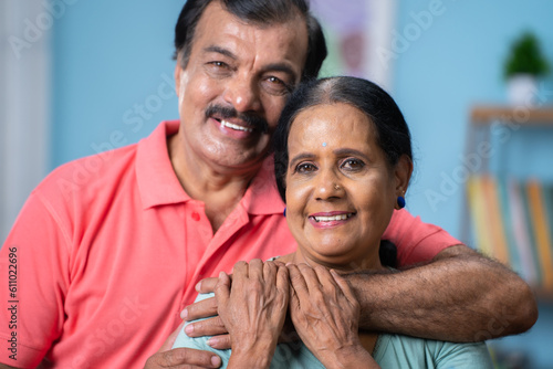Close up head shot of happy indian senior couple embracing each other by looking at camera at home - concept of relationship, family bonding and relaxation