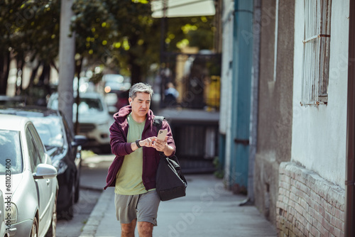 Middle aged man walking home from the gym on the sidewalk of a city street while using a smart phone