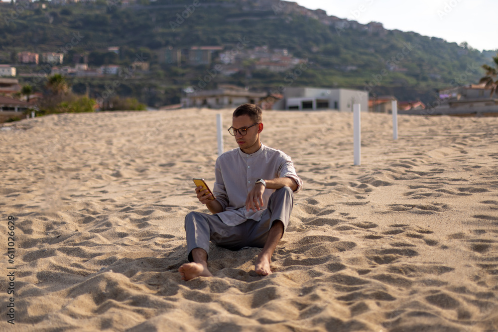 young caucasian man in a collarless shirt and dress pants, sitting on the sand and using the cell phone