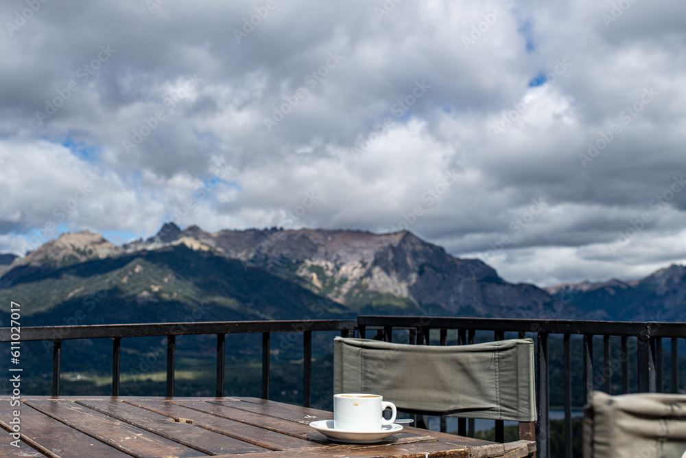 cup of coffee on a plate with a spoon on a wooden table next to a chair on a terrace overlooking the mountains from the cerro campanario in bariloche argentina