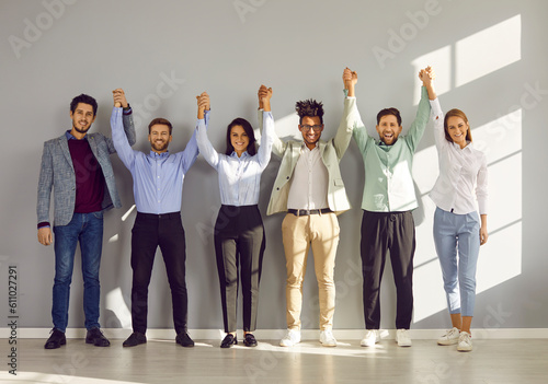 Happy confident business team holding hands raised together. Group of positive colleagues standing in line against wall in modern office, smiling at camera. Teamwork, team building and unity