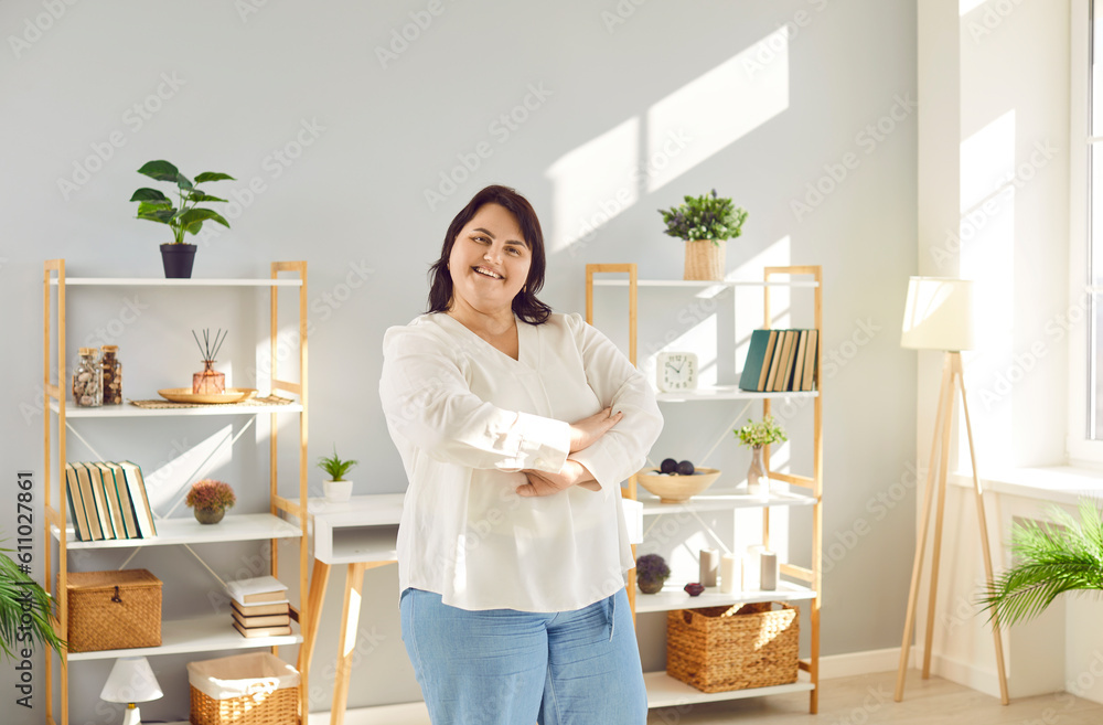 Portrait of a fat plus size brunette woman standing at home in the living room with crossed arms and smiling. Plump overweight cheerful girl looking positively at the camera. Body positive concept.