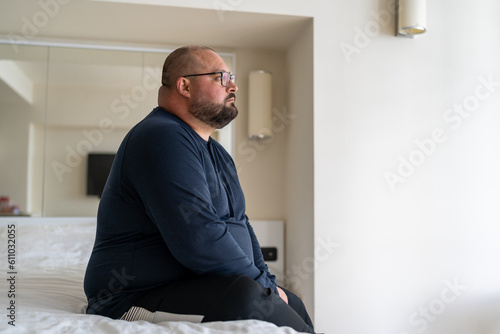 Bild auf Leinwand Sad fat lonely man sitting on bed in hotel and looking out window, thinking abou