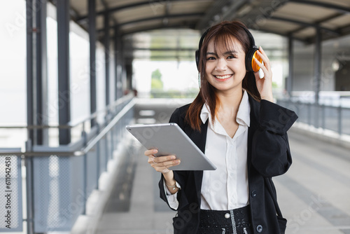 Attractive young woman in black suit holding tablet wearing headphone smiling looking a camera standing at public places. Lifestyle people technology concept. Female student outside university.