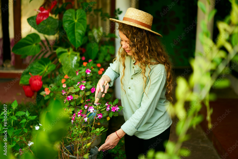 Young woman gardener in straw hat holding hand shovel taking care of potted plants. Junior caucasian female smiles standing in her little garden planting flowers in pots. Gardening and farming concept