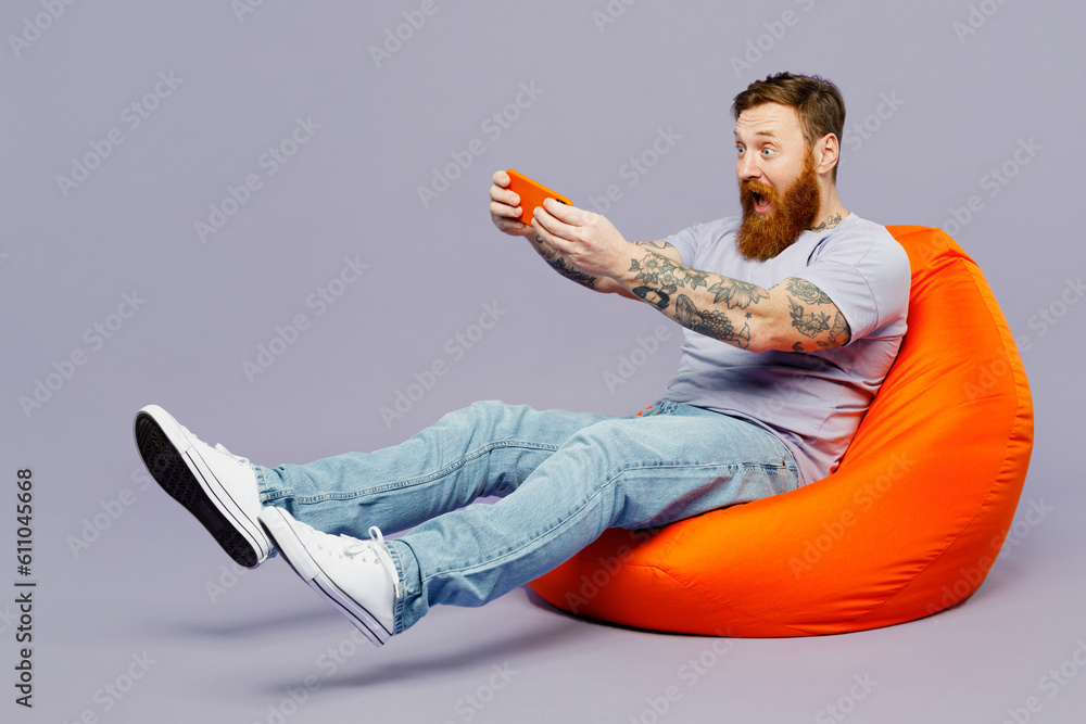 Full body young redhead man wear violet t-shirt casual clothes sit in bag chair use play racing app on mobile cell phone hold gadget smartphone for pc video games isolated on plain purple background. Full body young redhead man wear violet t-shirt casual clothes sit in bag chair use play racing app on mobile cell phone hold gadget smartphone for pc video games isolated on plain purple background.