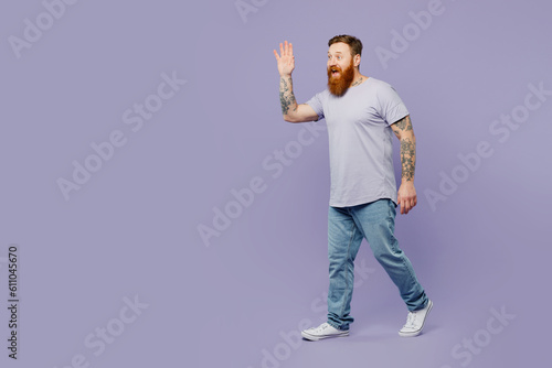 Full body side view young redhead bearded man he wearing violet t-shirt casual clothes walk go stroll waving hand isolated on plain pastel light purple background studio portrait. Lifestyle concept.